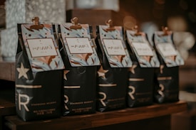 Five packages of Starbucks Reserve coffee with Hawaiian labels are neatly lined up on a wooden surface. The packaging features a dark design with stylized stars and the letter 'R'. Each package has a clip holding a colorful, tropical-themed label that reads 'Hawaii'. The background is softly blurred, creating a cozy and warm ambiance.