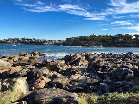 A rocky shoreline opens to a tranquil body of water with gentle waves. In the distance, a coastal community is visible with various buildings nestled along a green, tree-lined hillside under a vibrant blue sky with scattered wispy clouds.