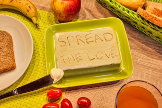 A block of butter with the words 'SPREAD THE LOVE' carved into it is placed on a green plate. Surrounding the butter are slices of bread, a banana, an apple, cherry tomatoes, and a glass of orange juice. These items are arranged on a wooden table with a green woven placemat.