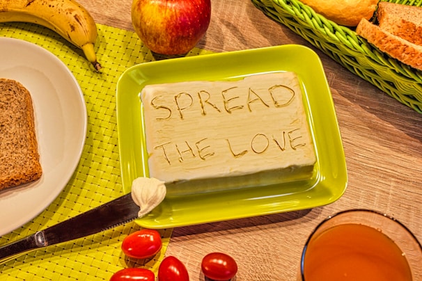 A block of butter with the words 'SPREAD THE LOVE' carved into it is placed on a green plate. Surrounding the butter are slices of bread, a banana, an apple, cherry tomatoes, and a glass of orange juice. These items are arranged on a wooden table with a green woven placemat.
