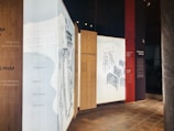 The image features a series of vertical panels displayed in an exhibition setting, with text and illustrations. The panels have a modern design, utilizing wooden textures and light backgrounds to highlight the text. Some panels contain drawings, including elements resembling ladders and chairs.