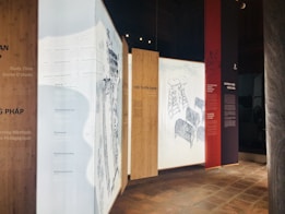 The image features a series of vertical panels displayed in an exhibition setting, with text and illustrations. The panels have a modern design, utilizing wooden textures and light backgrounds to highlight the text. Some panels contain drawings, including elements resembling ladders and chairs.