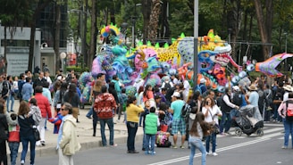 A street scene with a large crowd gathered around vibrant, intricately decorated, colorful sculptures resembling fantastical creatures. The sculptures are adorned with bright patterns and adornments, creating a festive atmosphere. People of all ages are observing and photographing the scene, while some are walking by with strollers or chatting. Trees and buildings in the background suggest an urban setting.