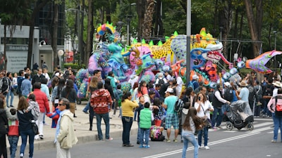 A street scene with a large crowd gathered around vibrant, intricately decorated, colorful sculptures resembling fantastical creatures. The sculptures are adorned with bright patterns and adornments, creating a festive atmosphere. People of all ages are observing and photographing the scene, while some are walking by with strollers or chatting. Trees and buildings in the background suggest an urban setting.