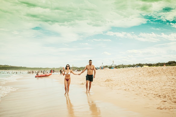 A couple walks hand in hand along a sandy beach. The woman is wearing a bikini and sunglasses, while the man is in swim shorts. The sky is partly cloudy, and people are scattered along the shoreline, some near a red inflatable boat.