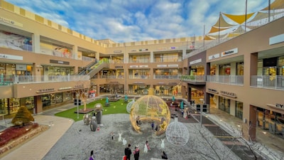 A multi-level shopping complex where the main courtyard features whimsical decorations including large ornamental balls with star patterns and artistic sculptures. The area is surrounded by various retail shops with signage visible for brands and stores. People are casually walking and enjoying the outdoor ambience, while overhead, a partly cloudy sky adds to the open atmosphere.