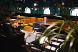 An elegant dining area with plush, gold and teal upholstered chairs surrounding dark wooden tables. The walls are adorned with vibrant, botanical wallpaper and round mirrors that enhance the room's sophisticated ambiance. Soft, spherical light fixtures hang overhead, while leafy plants add a touch of greenery.