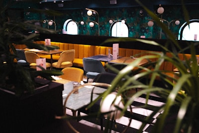 An elegant dining area with plush, gold and teal upholstered chairs surrounding dark wooden tables. The walls are adorned with vibrant, botanical wallpaper and round mirrors that enhance the room's sophisticated ambiance. Soft, spherical light fixtures hang overhead, while leafy plants add a touch of greenery.