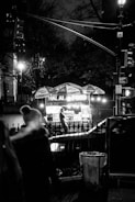 A nighttime scene featuring a street food cart with glowing lights, serving halal food. The cart is stationed on a city sidewalk, with a vendor visible inside. There are people passing by, and the setting is dimly lit, with streetlights and trees in the background.