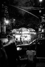 A nighttime scene featuring a street food cart with glowing lights, serving halal food. The cart is stationed on a city sidewalk, with a vendor visible inside. There are people passing by, and the setting is dimly lit, with streetlights and trees in the background.
