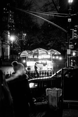 A nighttime scene featuring a street food cart with glowing lights, serving halal food. The cart is stationed on a city sidewalk, with a vendor visible inside. There are people passing by, and the setting is dimly lit, with streetlights and trees in the background.