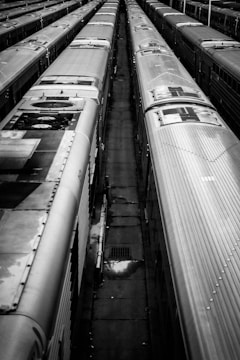 Multiple metallic train carriages are aligned in parallel tracks, viewed from above. The scene is monochromatic, highlighting the symmetrical rows and the industrial feel. The ground between the tracks is dark and appears slightly wet in areas, with the occasional small object scattered around.
