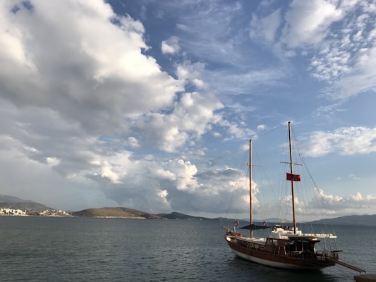 A wooden sailboat floats on a calm body of water, with large, fluffy clouds scattered across the sky. The horizon features distant hills and a small town along the shoreline. The boat is equipped with two tall masts and a red flag.