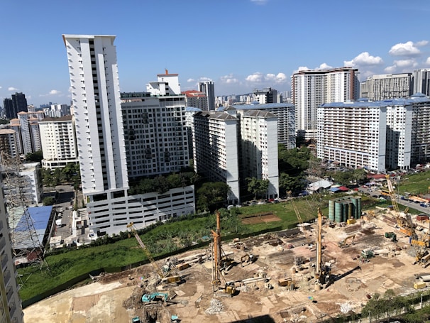 A large construction site is in the foreground with cranes and heavy machinery actively working on the site. Several high-rise residential buildings with repetitive architectural patterns dominate the background skyline against a clear blue sky. Scattered clouds add to the depth of the urban setting.