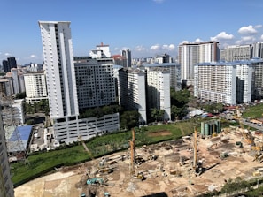 A large construction site is in the foreground with cranes and heavy machinery actively working on the site. Several high-rise residential buildings with repetitive architectural patterns dominate the background skyline against a clear blue sky. Scattered clouds add to the depth of the urban setting.