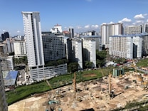 A large construction site is in the foreground with cranes and heavy machinery actively working on the site. Several high-rise residential buildings with repetitive architectural patterns dominate the background skyline against a clear blue sky. Scattered clouds add to the depth of the urban setting.
