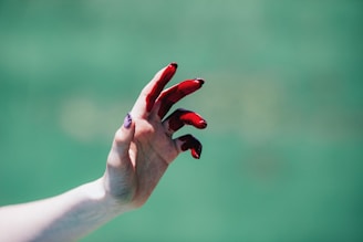 A hand is seen against a blurred green background. The fingers are partially covered in a red substance, possibly paint or a similar material. The lighting highlights the texture of the skin and the glossy nature of the red substance.