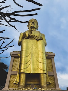 A large golden statue of a seated figure is prominently displayed against a backdrop of a cloudy sky. The statue exhibits intricate details in its drapery and posture. Bare branches of a tree frame the left side, adding contrast to the scene.