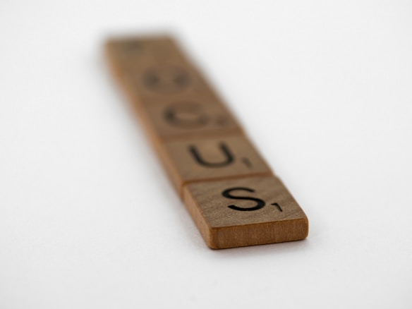 Wooden Scrabble tiles arranged to spell the word 'FOCUS' on a plain background. The tiles get progressively blurrier towards the top, emphasizing the foreground tiles.