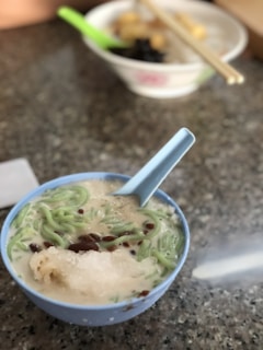 A bowl containing a dessert mixture of ingredients such as green jelly-like noodles, red beans, and a scoop of ice in coconut milk, accompanied by a light blue spoon. In the background, there's another bowl with chopsticks and food items, suggesting a setting in a casual dining environment.