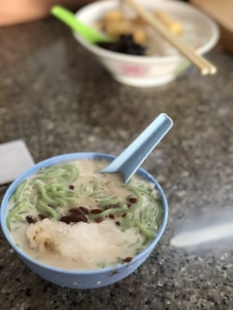 A bowl containing a dessert mixture of ingredients such as green jelly-like noodles, red beans, and a scoop of ice in coconut milk, accompanied by a light blue spoon. In the background, there's another bowl with chopsticks and food items, suggesting a setting in a casual dining environment.