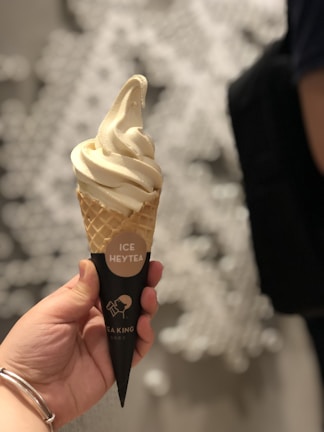 A hand holding a soft-serve ice cream cone with a light beige swirl. The cone is wrapped in a black paper wrapper with the text 'ICE HEYTEA' and a logo. The background is softly blurred, with abstract white shapes.