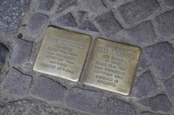 Two brass plaques are embedded in a cobblestone path. They bear inscriptions commemorating Jakob and Felli Bergoffen, detailing their escape from Prague in 1938, deportation from France in 1942, and eventual murder in Auschwitz.