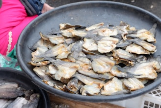 A large pile of dried fish is placed in a black plastic basket. The fish have a light, golden-brown coloration and appear to be arranged in a casual, overlapping fashion. A person in vibrant pink and blue clothing is visible partially in the background.