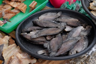 A black circular tray is filled with several whole dried fish stacked on top of each other. To the side, a green tray contains various cuts of dried fish displayed in an orderly fashion. The background shows additional dried fish and a hint of colorful materials.