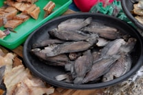 A black circular tray is filled with several whole dried fish stacked on top of each other. To the side, a green tray contains various cuts of dried fish displayed in an orderly fashion. The background shows additional dried fish and a hint of colorful materials.