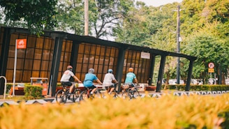 Four cyclists ride along a designated bike pathway next to a modern bus stop shelter. The area is surrounded by lush green trees and bushes, creating a natural and serene environment. There are traffic signs indicating rules for both cyclists and vehicles.