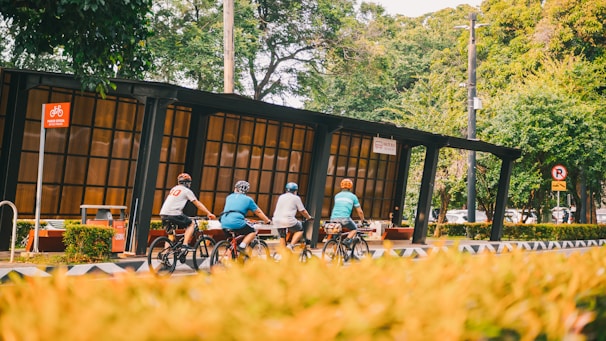 Four cyclists ride along a designated bike pathway next to a modern bus stop shelter. The area is surrounded by lush green trees and bushes, creating a natural and serene environment. There are traffic signs indicating rules for both cyclists and vehicles.