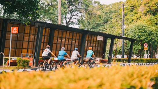 Four cyclists ride along a designated bike pathway next to a modern bus stop shelter. The area is surrounded by lush green trees and bushes, creating a natural and serene environment. There are traffic signs indicating rules for both cyclists and vehicles.