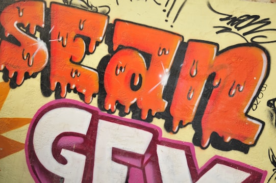 Colorful graffiti artwork featuring the word 'SEAN' in large, orange, gradient letters with a dripping effect. The text is outlined in black with some white highlights. Below 'SEAN,' there are white and purple block letters. The background is light yellow, with additional black and purple graffiti tags and art surrounding the main text.