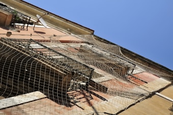The image features a building facade with a beige-pink exterior, accented by open windows and balconies. A satellite dish is mounted near the top of the structure. Safety nets are draped over parts of the building, possibly a precaution for maintenance work or structural integrity.