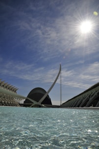 An architectural scene featuring modern structures with futuristic designs reflected in a pool of water under a bright sky with the sun shining prominently.