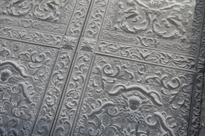 Intricately designed metal ceiling tiles featuring elaborate floral and vine patterns. The tiles are arranged in a grid, creating a repetitive motif across the surface. The relief designs give the surface a textured appearance.