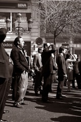 A group of people stands on a city street, dressed in formal and casual attire. They appear to be looking towards something of interest off-frame. The background includes a bank sign and a bare tree, suggesting a business district. The photo is in black and white, emphasizing the contrast and textures.