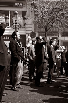 A group of people stands on a city street, dressed in formal and casual attire. They appear to be looking towards something of interest off-frame. The background includes a bank sign and a bare tree, suggesting a business district. The photo is in black and white, emphasizing the contrast and textures.