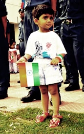 A young child is holding the Indian flag and standing on a grassy area, surrounded by adults dressed in dark uniforms. The child is wearing a white t-shirt with a print on it, white shorts, and red sandals. In the background, there are other people, including children, some of whom are also holding flags. The scene appears to be a public event or a parade.