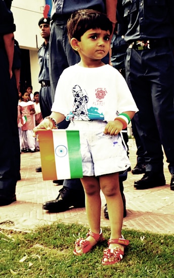 A young child is holding the Indian flag and standing on a grassy area, surrounded by adults dressed in dark uniforms. The child is wearing a white t-shirt with a print on it, white shorts, and red sandals. In the background, there are other people, including children, some of whom are also holding flags. The scene appears to be a public event or a parade.