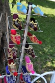 A display rack holds colorful, traditional embroidered slippers, featuring intricate gold patterns and vibrant pom-poms on the toes. The slippers are arranged vertically in pairs with a lush green lawn in the background.