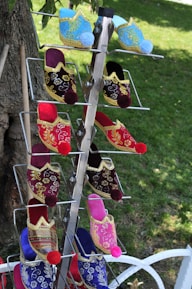 A display rack holds colorful, traditional embroidered slippers, featuring intricate gold patterns and vibrant pom-poms on the toes. The slippers are arranged vertically in pairs with a lush green lawn in the background.