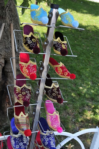 A display rack holds colorful, traditional embroidered slippers, featuring intricate gold patterns and vibrant pom-poms on the toes. The slippers are arranged vertically in pairs with a lush green lawn in the background.