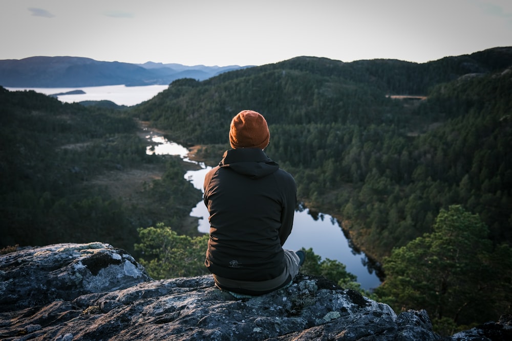 man in black hoodie sitting on rock looking at lake during daytime