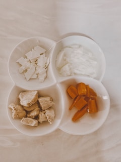 A white, three-sectioned dish contains various food items: chopped white cheese, a portion of white yogurt or cream, chunks of cooked chicken, and sliced orange carrots. The dish is set on a light marble surface.