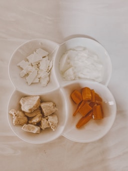 A white, three-sectioned dish contains various food items: chopped white cheese, a portion of white yogurt or cream, chunks of cooked chicken, and sliced orange carrots. The dish is set on a light marble surface.