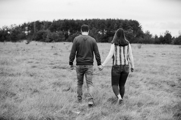 A couple is walking hand in hand across an open grassy field with a forest of trees in the background. The image is in black and white, emphasizing a sense of connection and nature. The couple is dressed casually, with the woman wearing a fur vest and the man in a sweater.