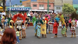 A group of people dressed in traditional, colorful indigenous costumes are participating in a street parade. The costumes feature elaborate feathered headdresses and ornate patterns. Children and adults are gathered, some holding musical instruments like drums. A pastel-colored building with a sign that reads 'PASTELERIA' is visible in the background.