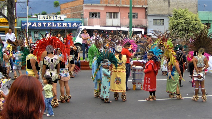 A group of people dressed in traditional, colorful indigenous costumes are participating in a street parade. The costumes feature elaborate feathered headdresses and ornate patterns. Children and adults are gathered, some holding musical instruments like drums. A pastel-colored building with a sign that reads 'PASTELERIA' is visible in the background.